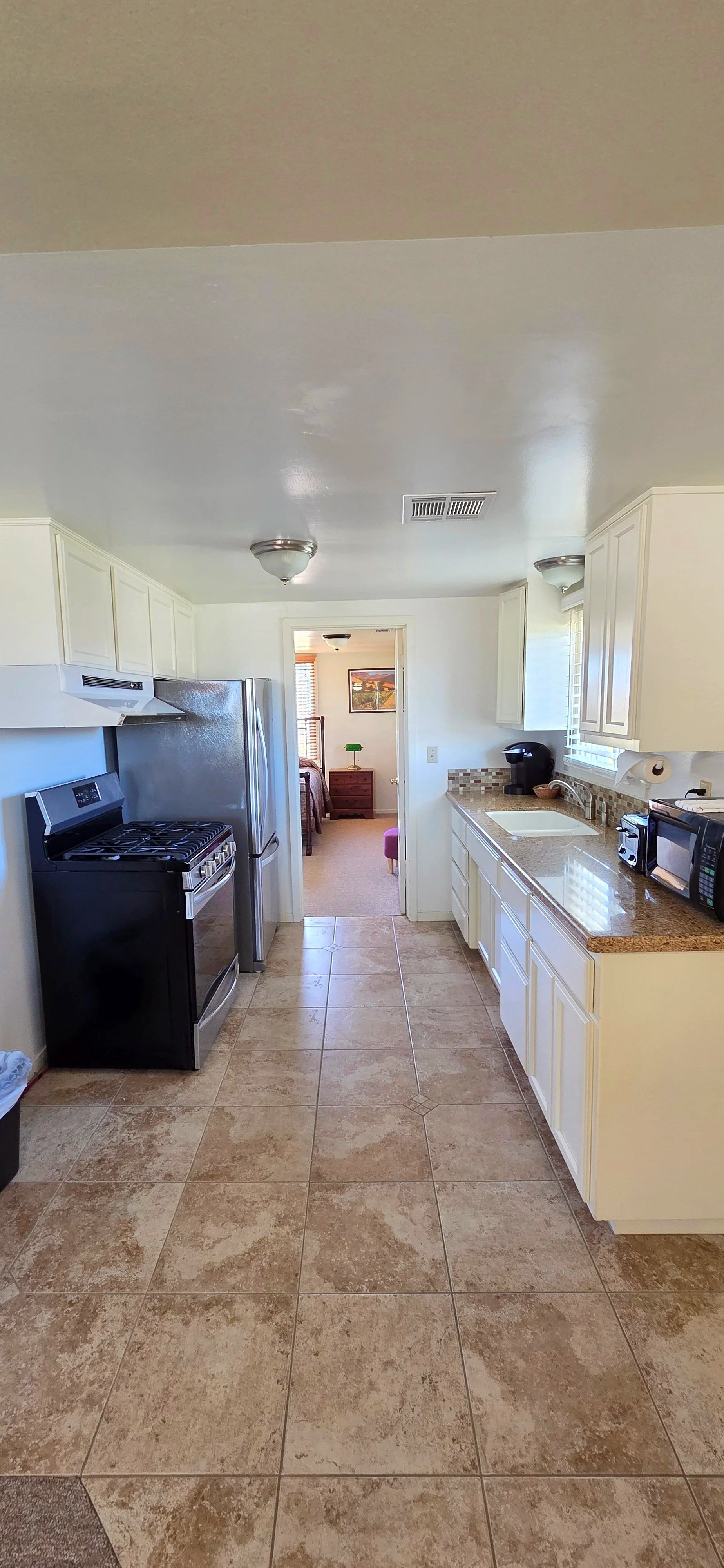 A kitchen with beige tile flooring, white cabinets, and stainless steel appliances including a refrigerator and stove. There is a black coffee maker, microwave, and a small window with blinds above the sink. A doorway leads to a bedroom with a bed, nightstand, and a window with blinds.