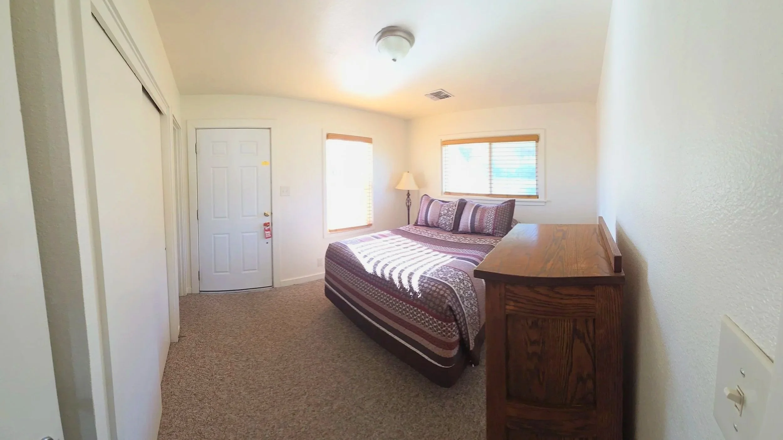 A bedroom with a bed, window blinds, a nightstand with a lamp, and a closet door.