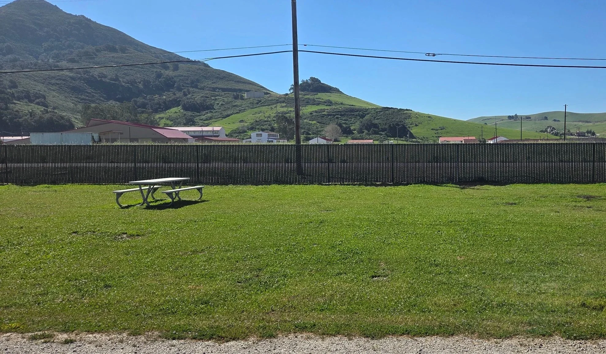 Open grassy field with a picnic table, black fence, hills and houses in the background, and a blue sky.