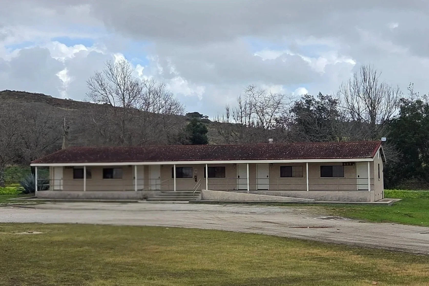 Single-story beige building with a red-tiled roof, several windows, a small staircase, and a wheel chair accessible ramp, set against a backdrop of leafless trees and cloudy sky.