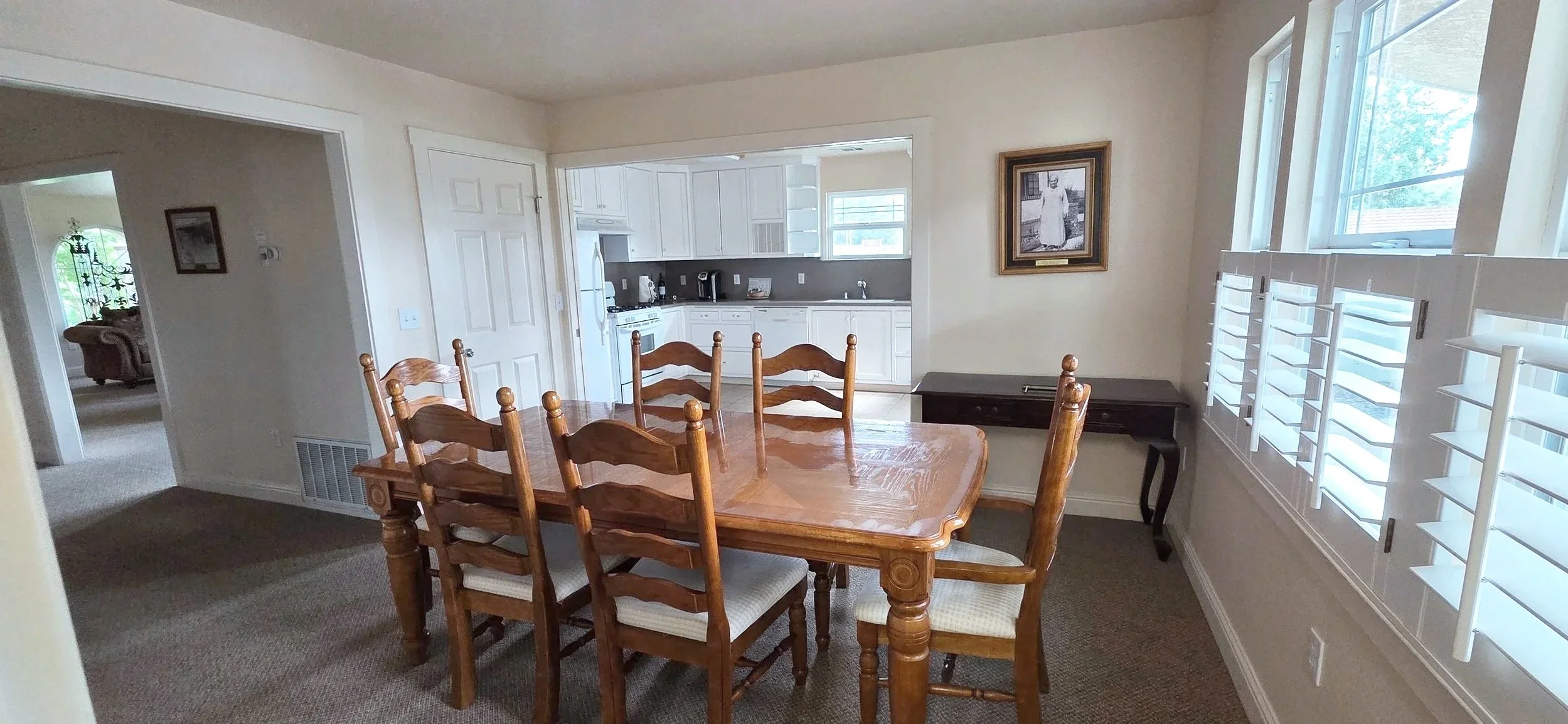 Dining room with wooden table and six chairs, large windows with shutters, and a view into the kitchen with white cabinets and appliances.