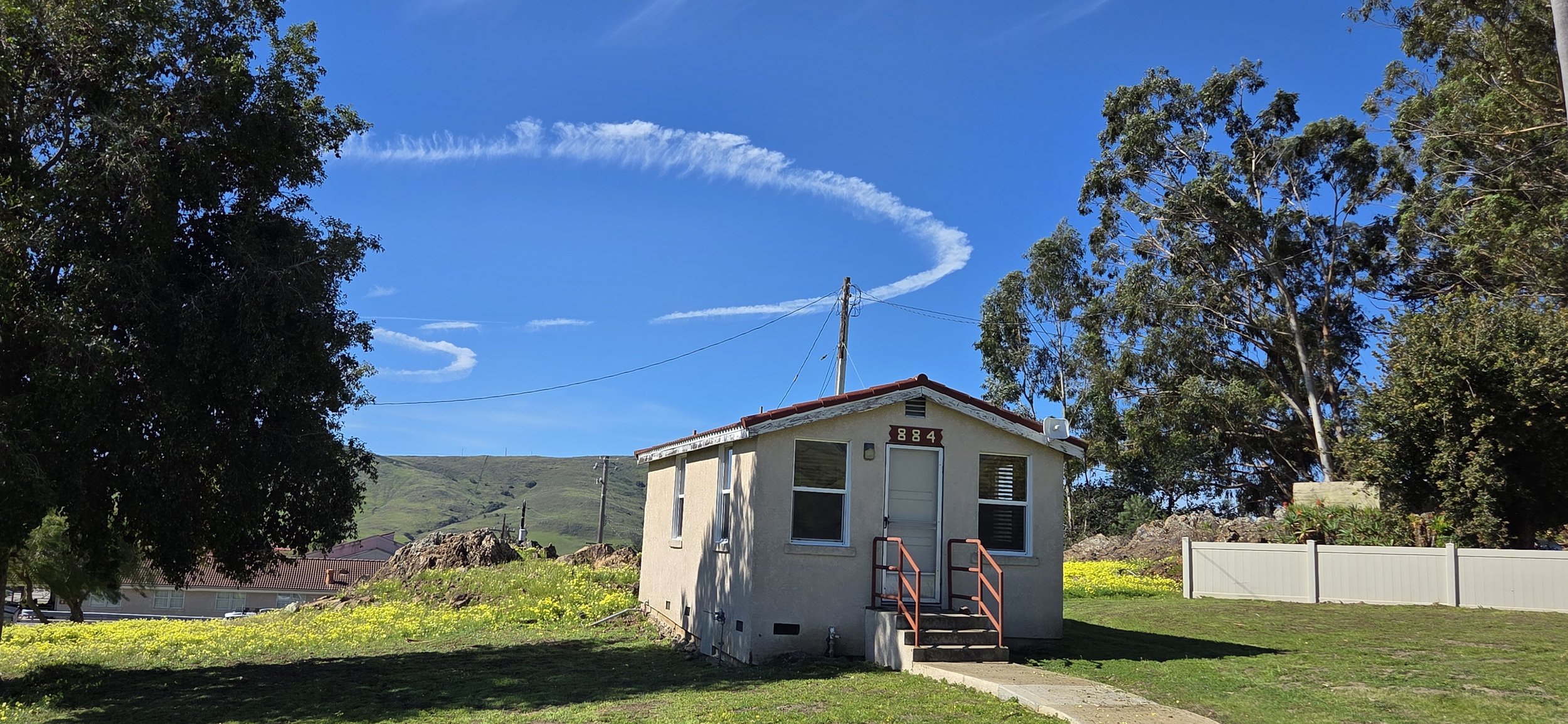 Small beige house with red roof and steps in a grassy yard, surrounded by trees with a blue sky and clouds in the background.