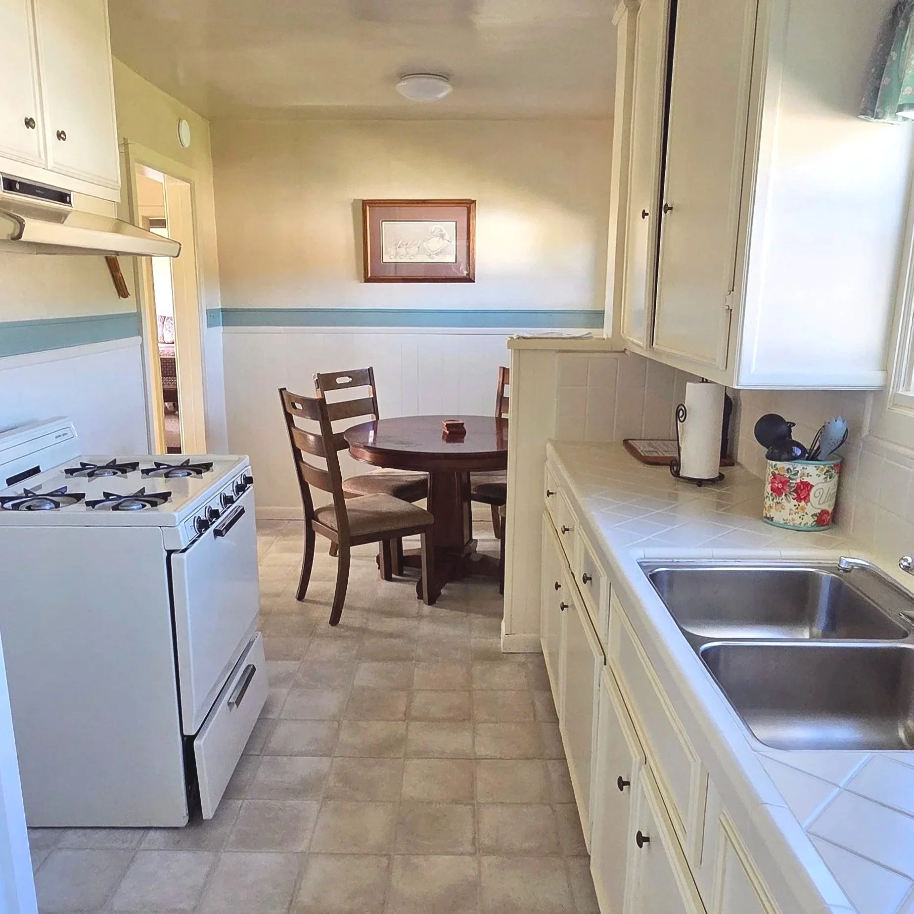 A kitchen with white cabinets, a double sink, a gas stove, a small round dining table with three wooden chairs, and a framed picture on the wall.