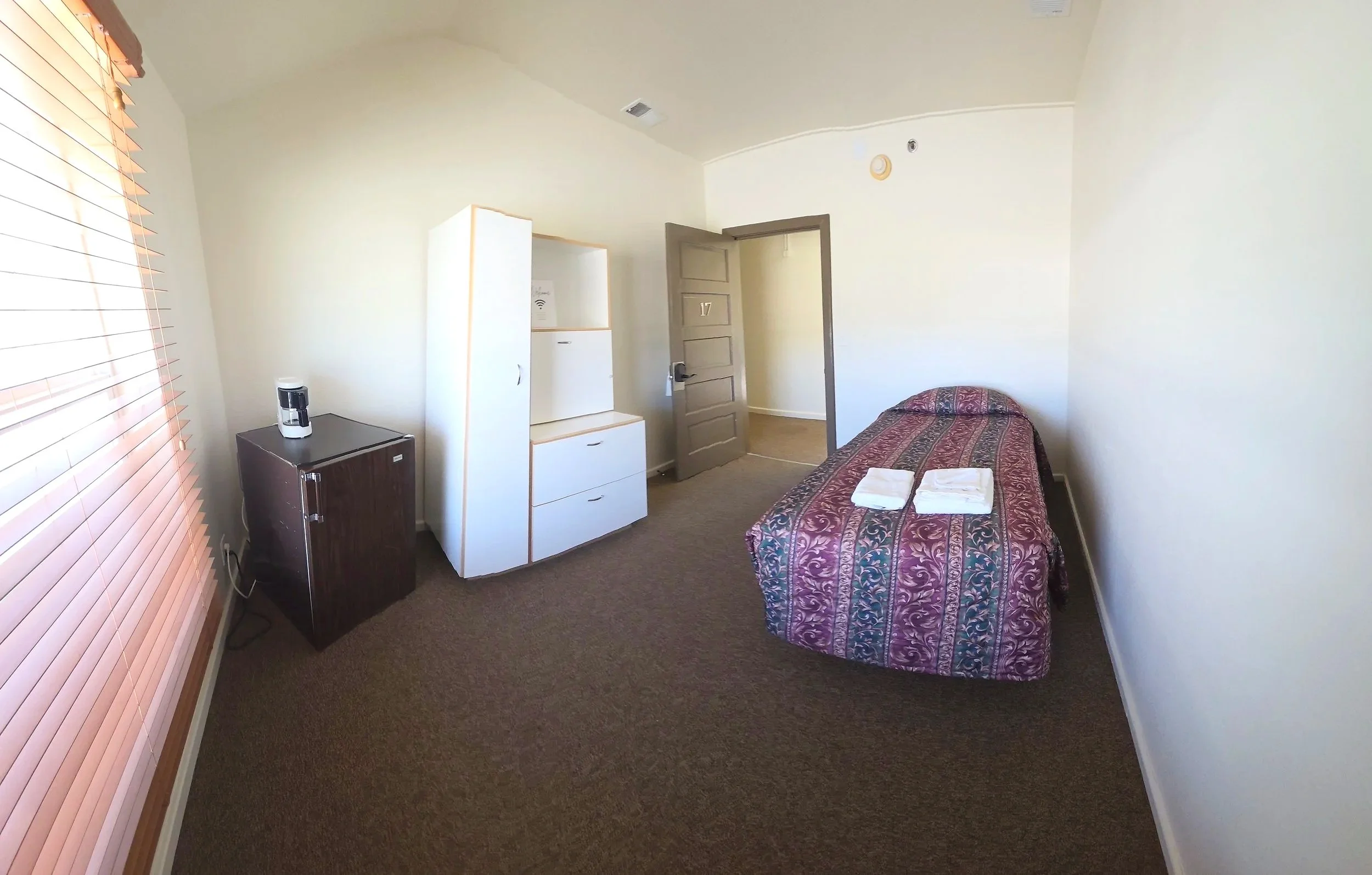 A small hotel room with a single bed, folded towels on the bed, a window with blinds, a small refrigerator, a white wardrobe, and an open door leading to a hallway.