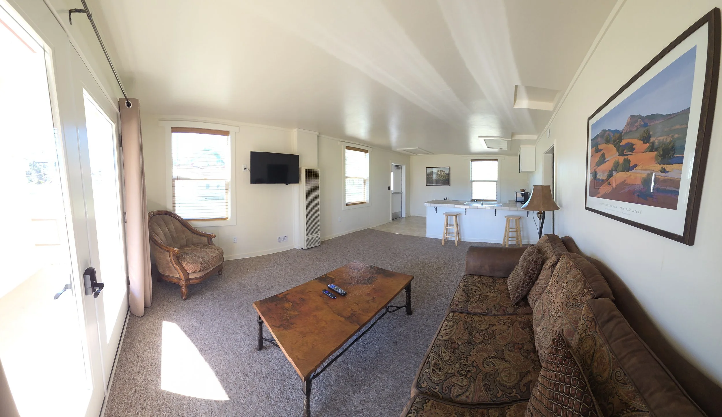 Living room with a brown sofa, armchair, wooden coffee table, wall-mounted TV, and framed landscape art, with windows and a sliding glass door.