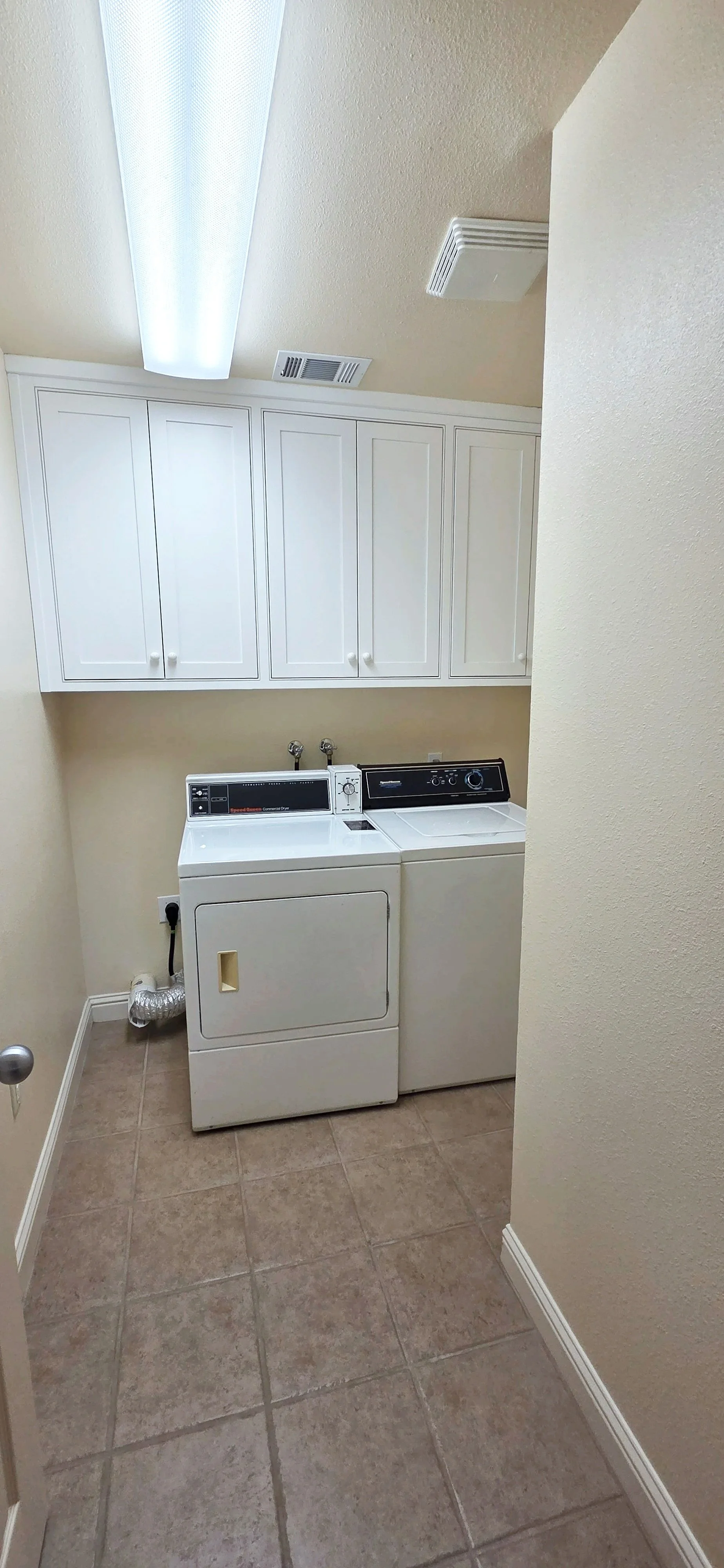 Laundry room with a washer and dryer, white cabinets above, beige walls, tile floor, and fluorescent ceiling light.