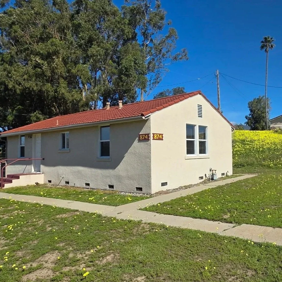 A light-colored house with a red tiled roof, two windows on the front, and a small set of stairs leading to the entrance, surrounded by a grassy lawn with scattered yellow flowers and a sidewalk.