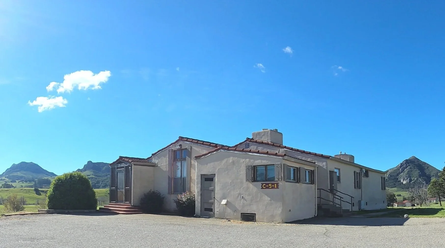A white, single-story building with a red tile roof situated in a rural landscape with green hills and mountains in the background. Clear blue sky with a few small clouds.