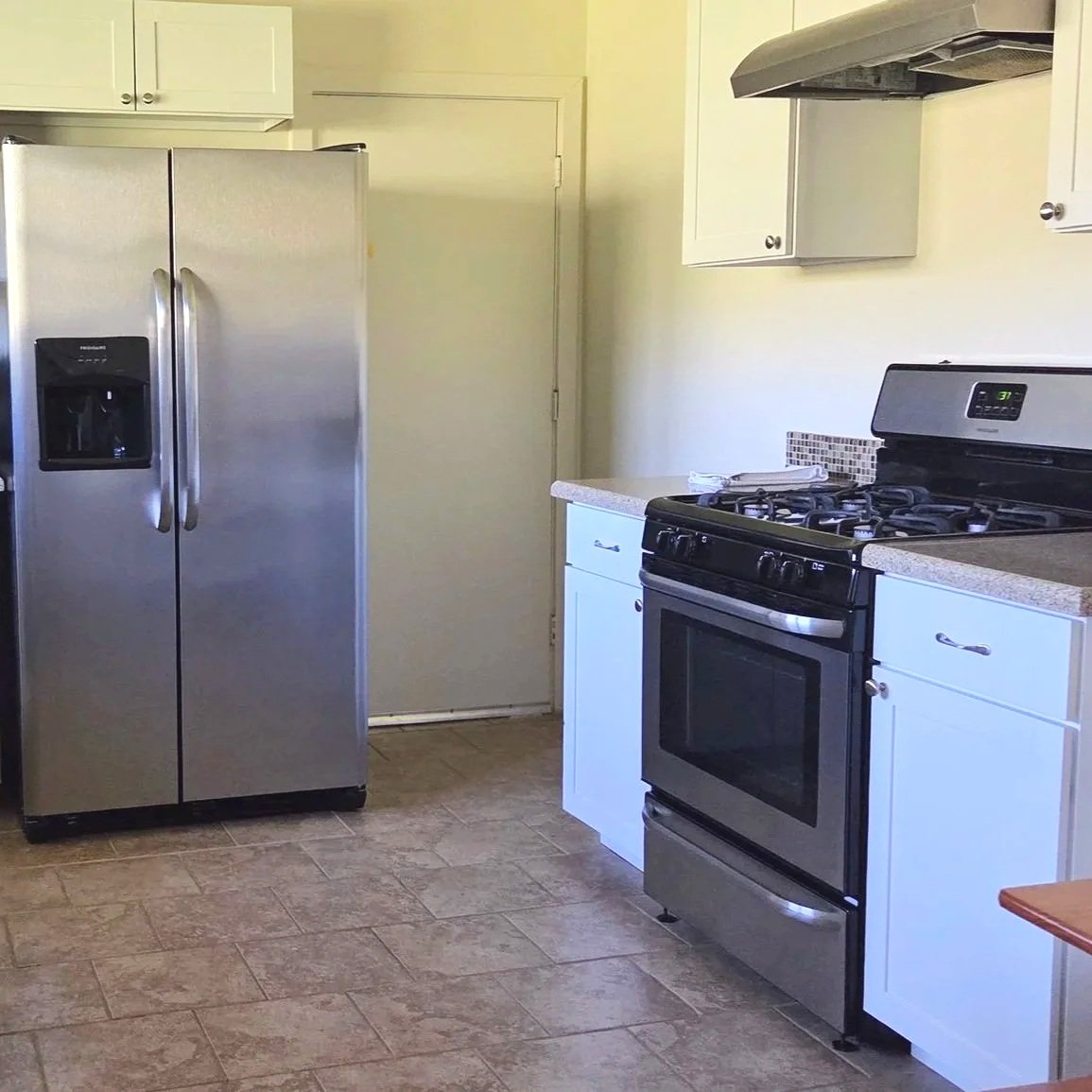 A kitchen with a stainless steel refrigerator on the left and a black oven with a gas stovetop on the right. White cabinets are above and below the countertops, and there is a tiled backsplash. The floor is tiled with a brownish pattern.