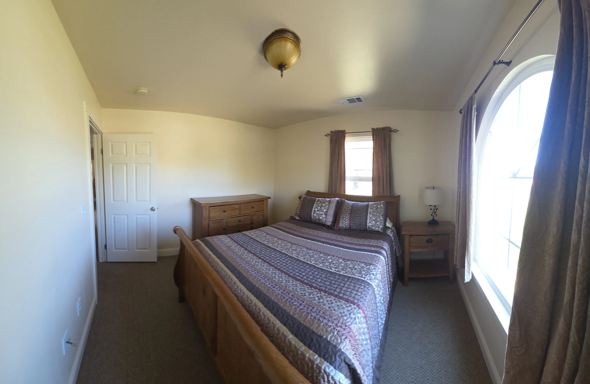 A bedroom with a wooden bed frame, striped quilt, matching pillows, two windows with brown curtains, a wooden dresser, a bedside table with a lamp, and beige walls.