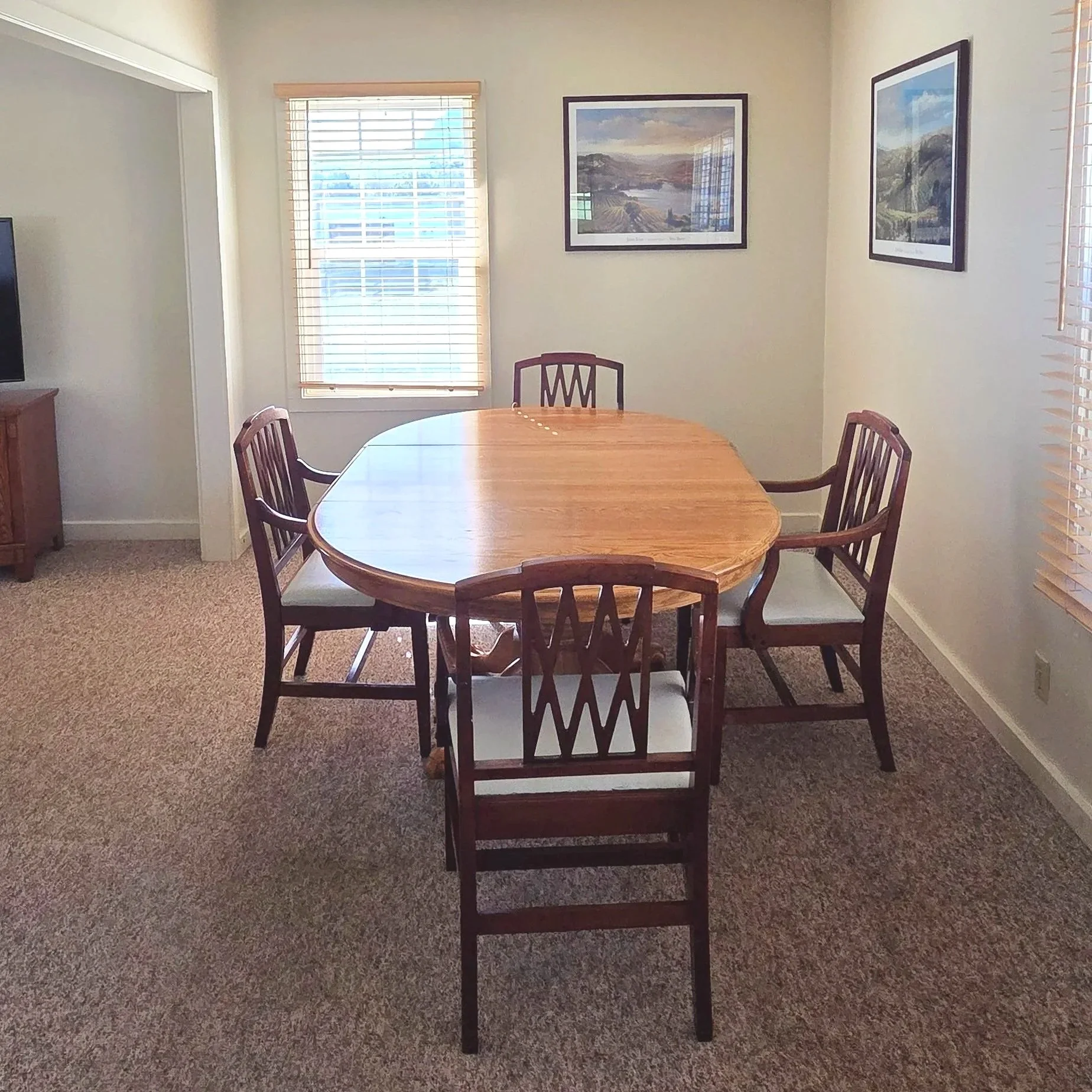 Dining room with a wooden oval table and five matching chairs with white cushions, multiple windows with blinds, and framed landscape pictures on the wall.