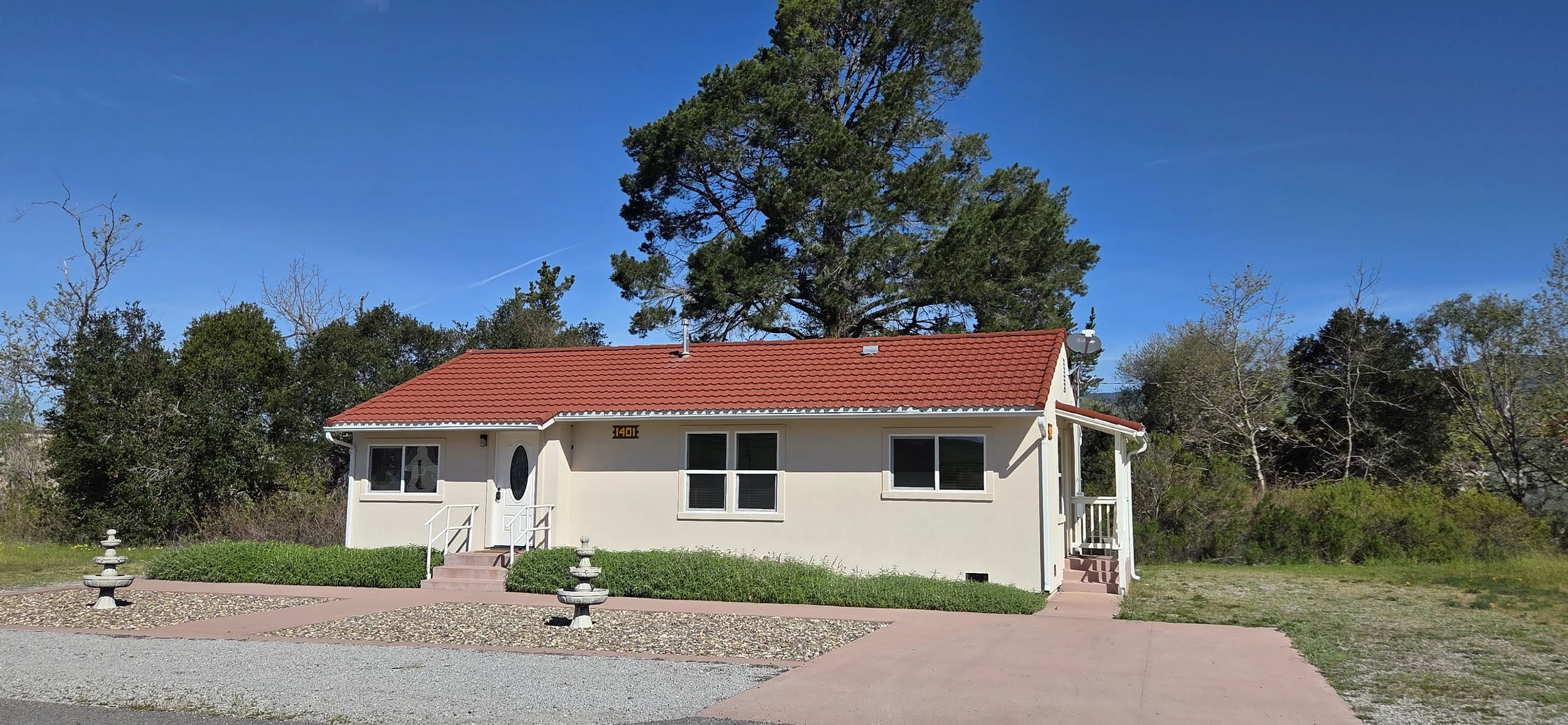 Single-story house with white exterior, red tiled roof, front steps, and small porch, situated in a semi-rural area with trees and a gravel driveway.