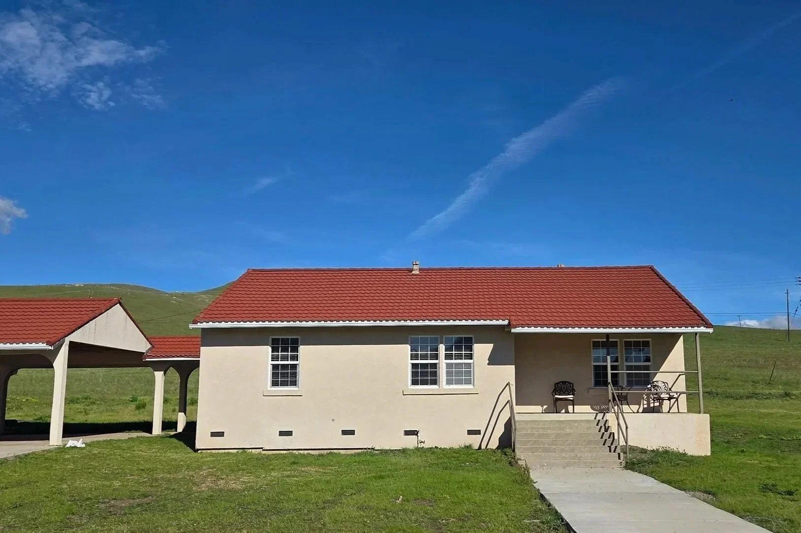 A house with a red tiled roof, light-colored walls, and a small front porch with chairs. Green grass and a hillside are in the background under a blue sky.