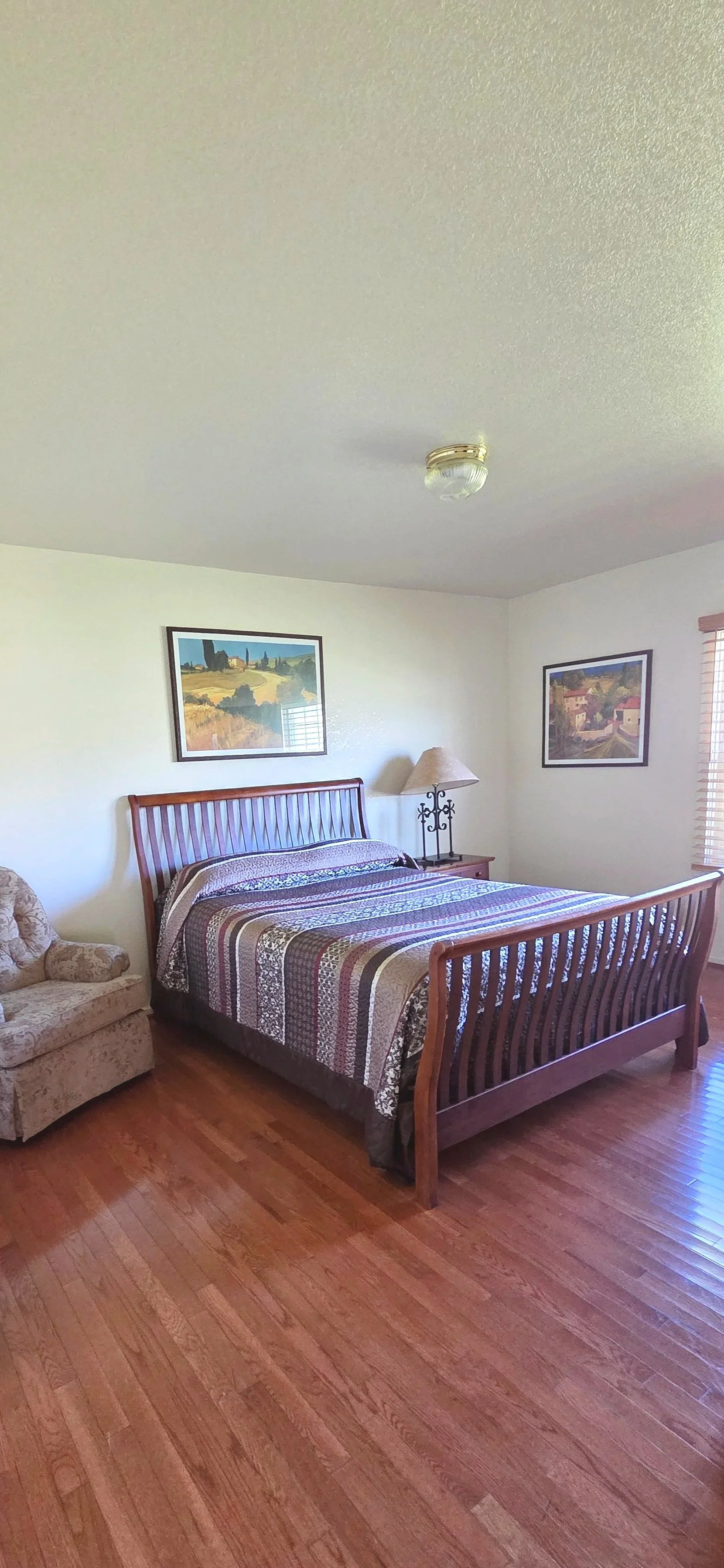 A bedroom with a wooden bed, a patterned bedspread, a beige armchair, a nightstand with a lamp, and framed artwork on the wall, with hardwood floors and a window with blinds.