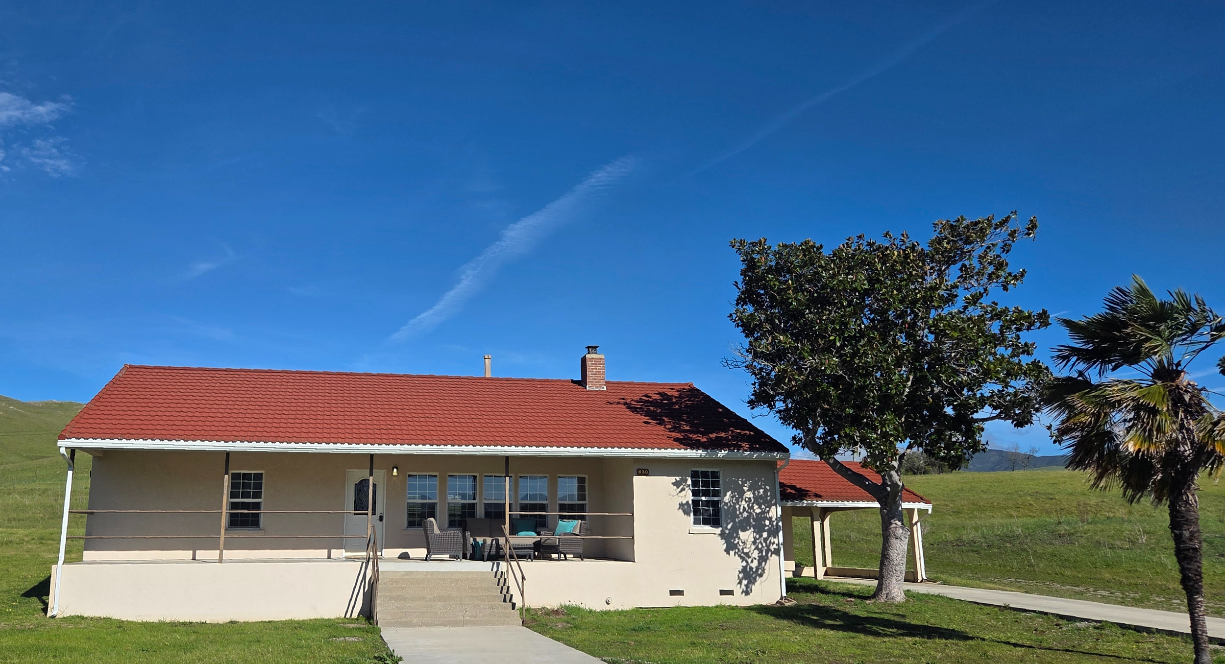 Single-story house with beige walls, red tile roof, front porch with outdoor furniture, surrounded by grassy land and trees, under a clear blue sky.