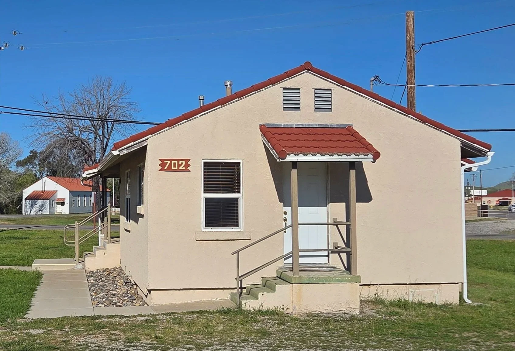 A small beige house with a red tile roof and steps leading up to the white front door, with a small porch and metal railing, set against a clear blue sky.