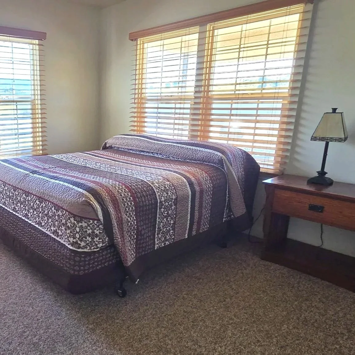 A bedroom with a bed, a wooden nightstand, and a lamp. The bed has a colorful quilt, and there are large windows with wooden blinds letting in natural light.