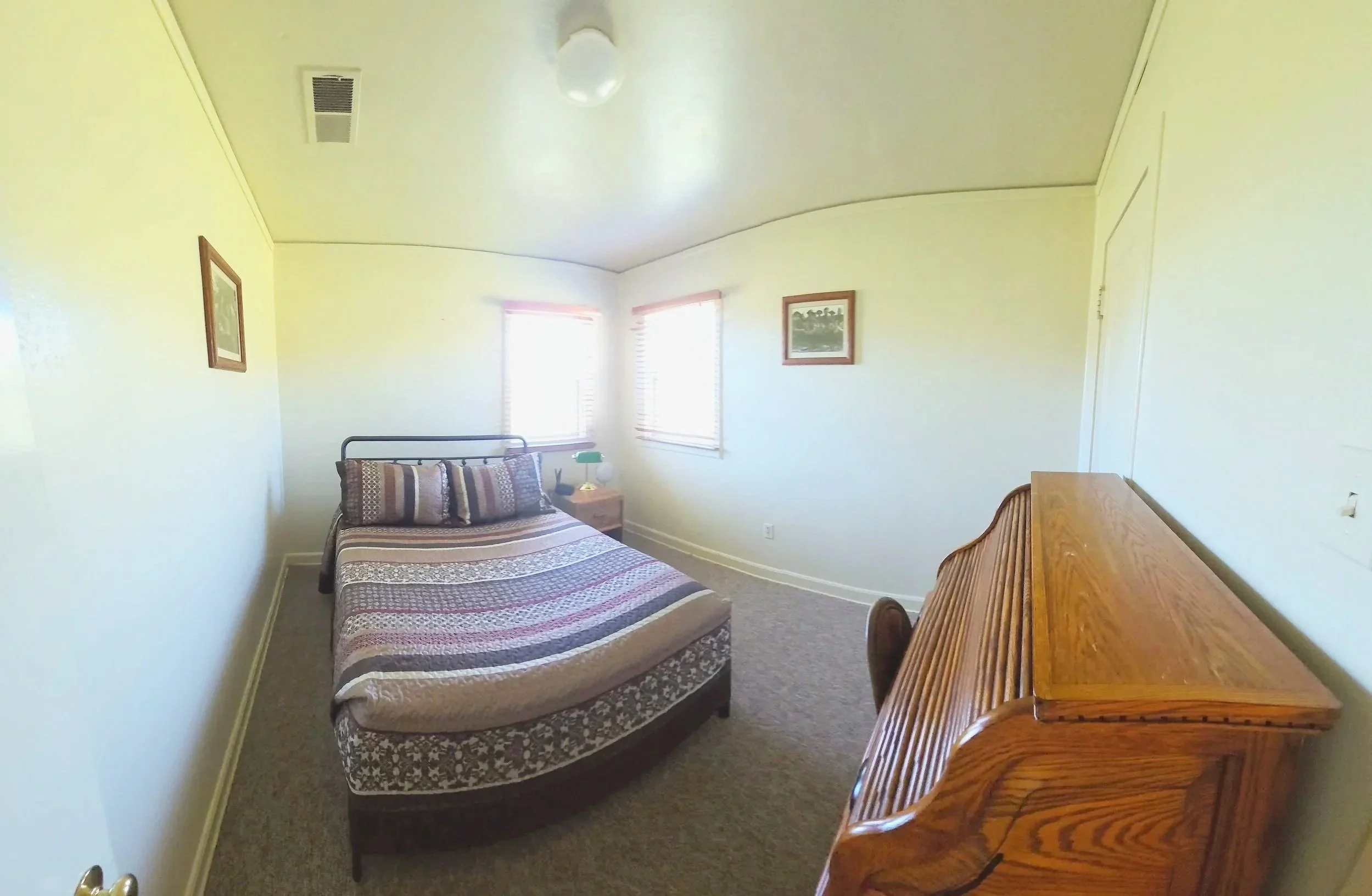 A small bedroom with a bed, nightstand, and a wooden desk, illuminated by sunlight from two windows with blinds, hanging pictures on the walls, and a ceiling light fixture.