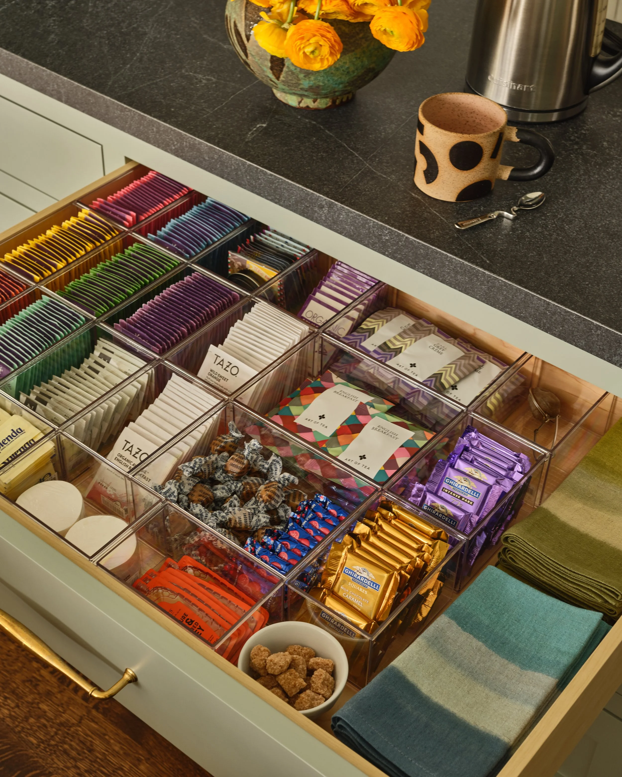 A drawer filled with various tea bags sorted by color, with a small bowl of brown sugar cubes and two rolled cloths on top. On the counter above, there is a vase with yellow flowers, a coffee mug, a kettle, and some napkins.