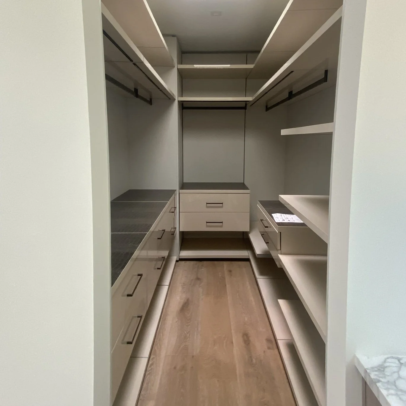 Empty walk-in closet with beige shelves and drawers, wooden flooring.