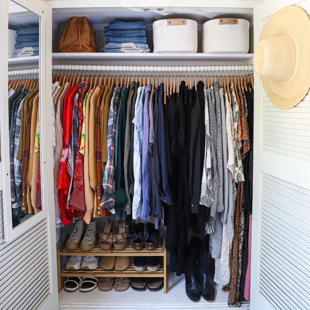 A closet with clothes hanging on a rod, shoes on shelves at the bottom, and folded clothes and storage boxes on the top shelf. A hat hangs on the right side.