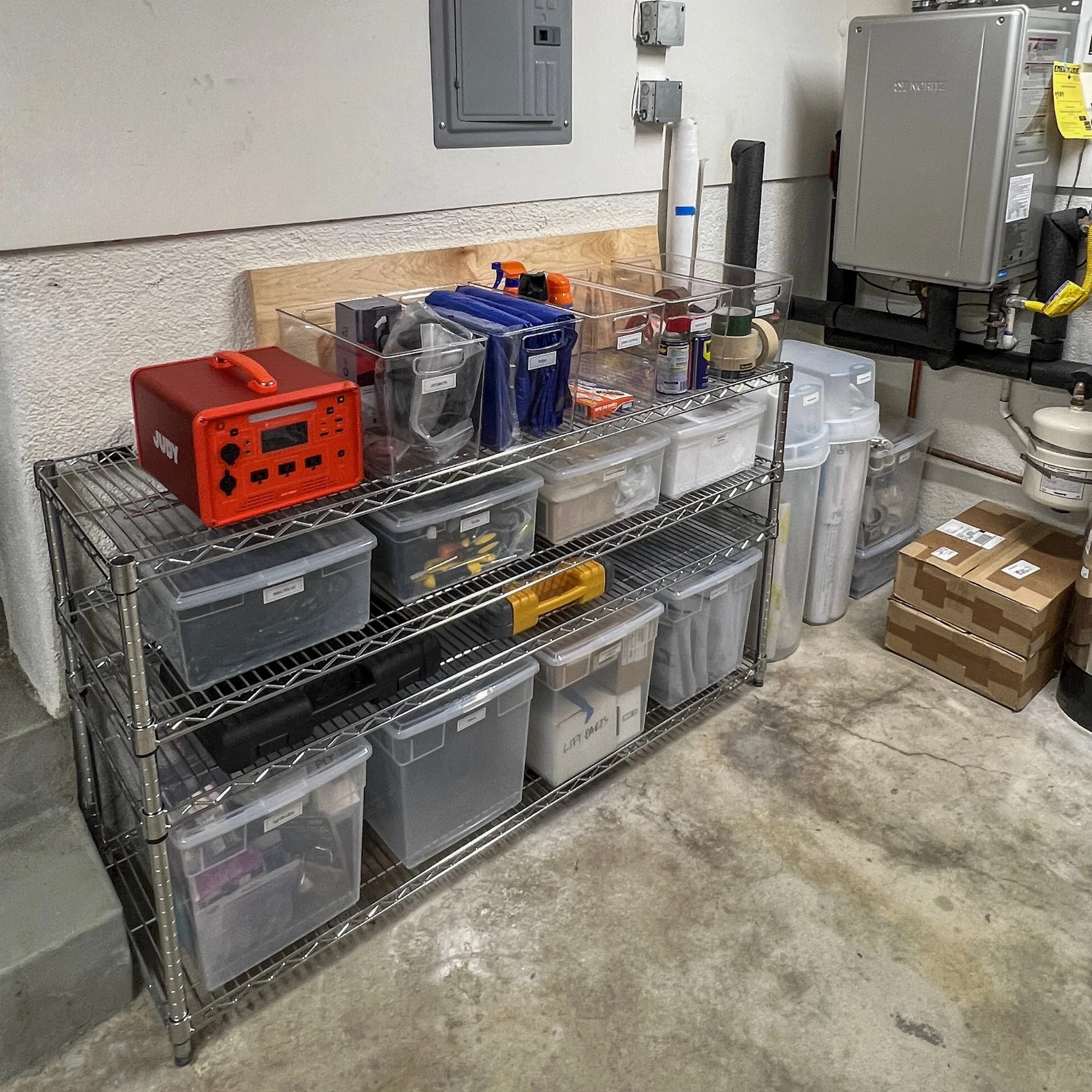 A metal shelving unit with clear plastic containers filled with tools and supplies, a red toolbox, and a cardboard box on the floor next to it in a utility room or workshop.