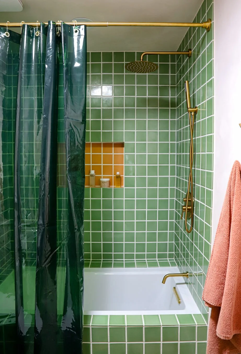 Green tiled shower with a black showerhead and a hand-held shower wand, gold fixtures, a built-in shower niche with toiletries, and a pink towel hanging on the side.
