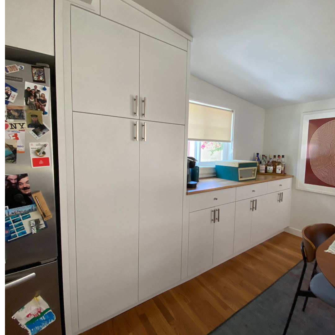 A kitchen with white cabinets, a wooden countertop, a window with a window shade, a coffee maker, a microwave, and bottles of alcohol on the counter. A stainless steel refrigerator with photos and magnets is visible on the left. There is a wooden tab