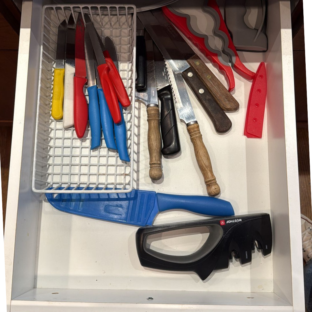 A drawer containing various kitchen knives with colorful handles, a serrated bread knife, a meat cleaver, a hacksaw, safety knives with red handles, a blue spatula, and a black safety cutter.