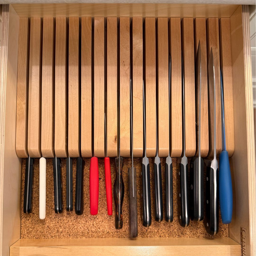 A wooden knife block with eleven knives of various sizes and handle colors stored inside, placed in a kitchen drawer.