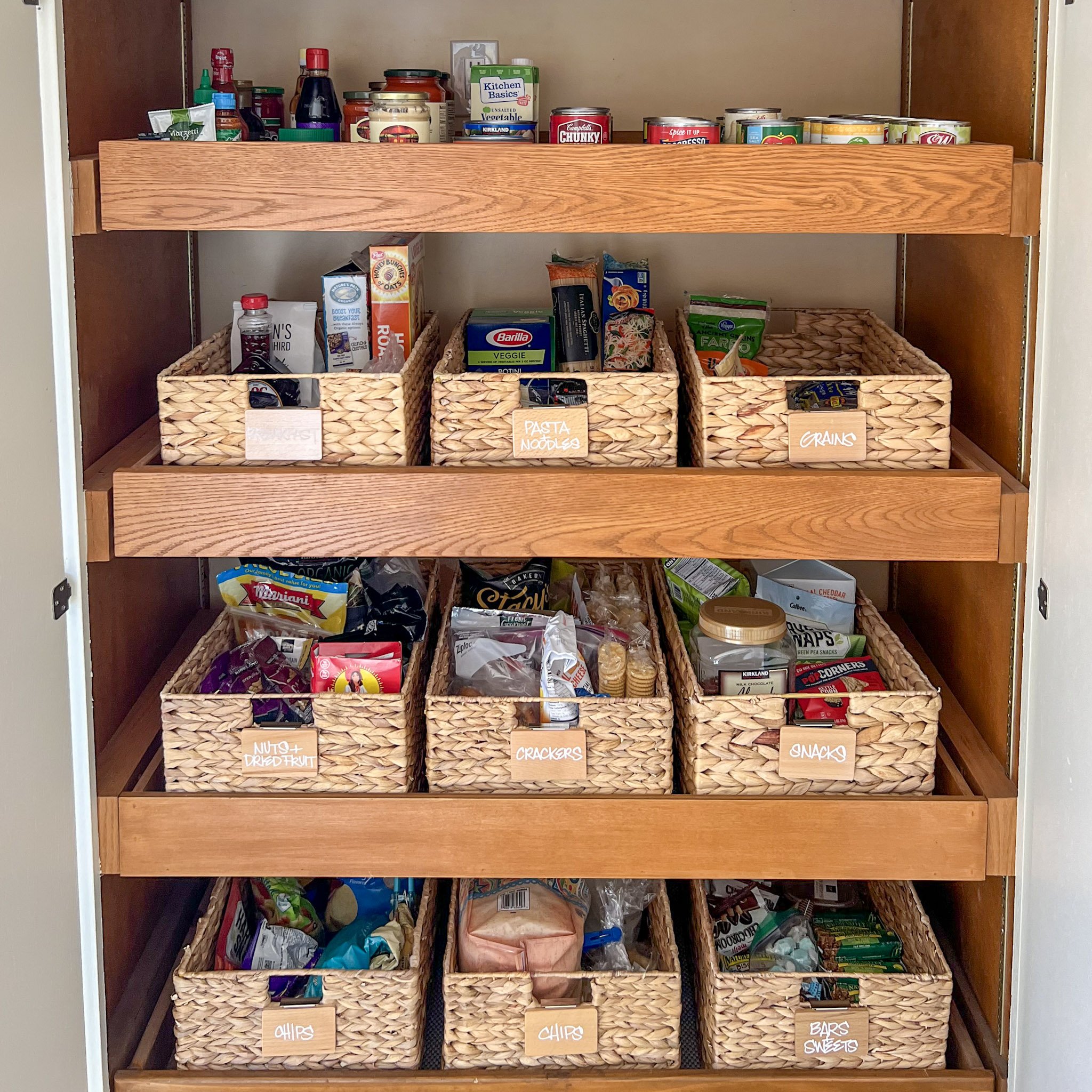 A wooden pantry shelf organized with four woven baskets labeled 'Nuts & Dried Fruit,' 'Crackers,' 'Chips,' and 'Bars & Sweets,' containing various snacks and packaged foods.