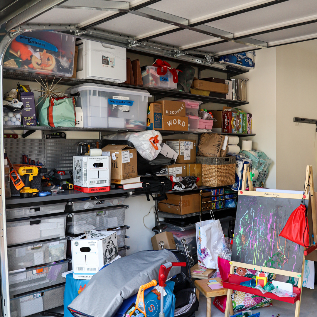 A cluttered garage with shelving unit filled with boxes, tools, toys, and art supplies, including a chalkboard with colorful drawings and a small wooden table with toys and supplies.