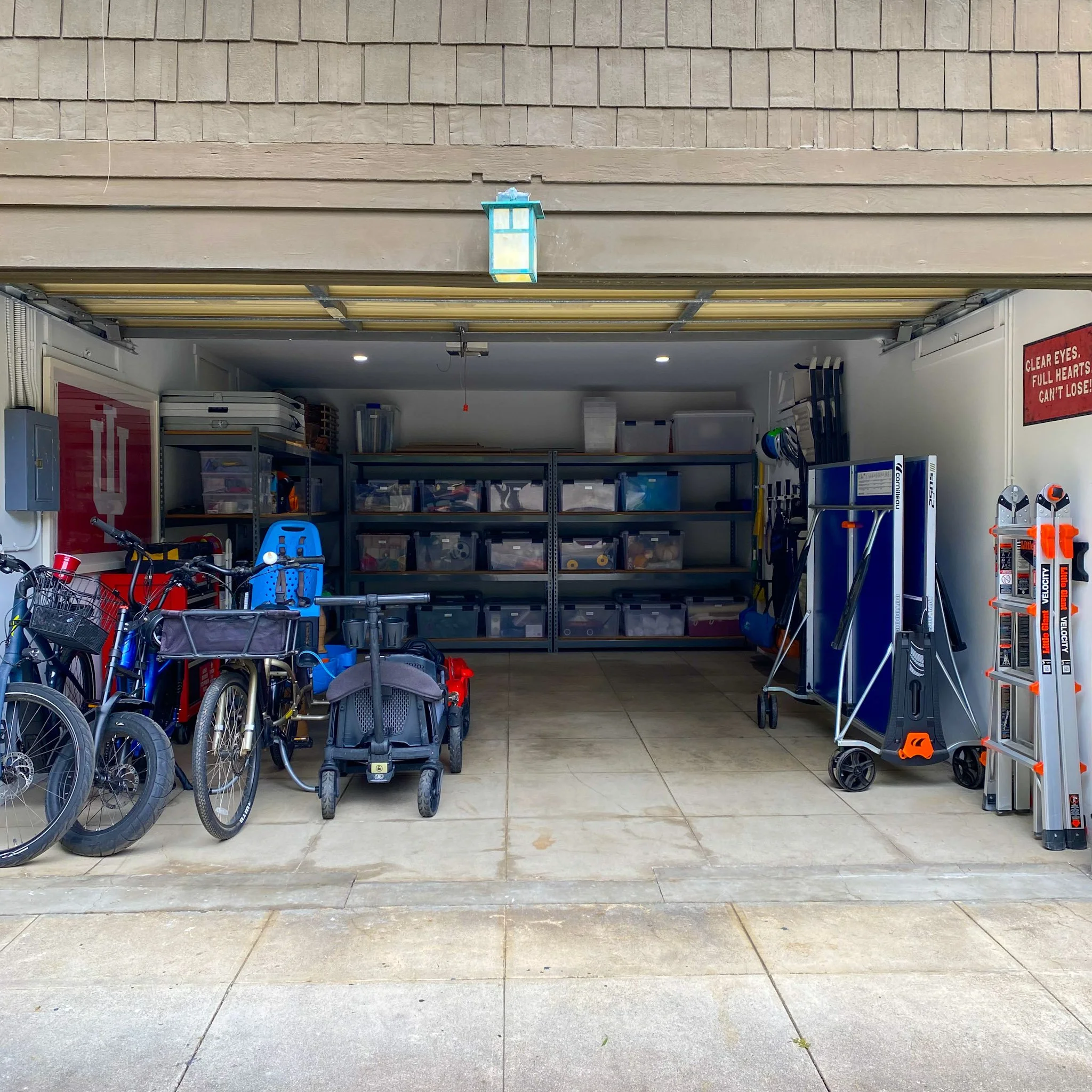 View of a garage with bicycles, storage bins, and tools inside. The garage door is open, showing a textured brown exterior wall.