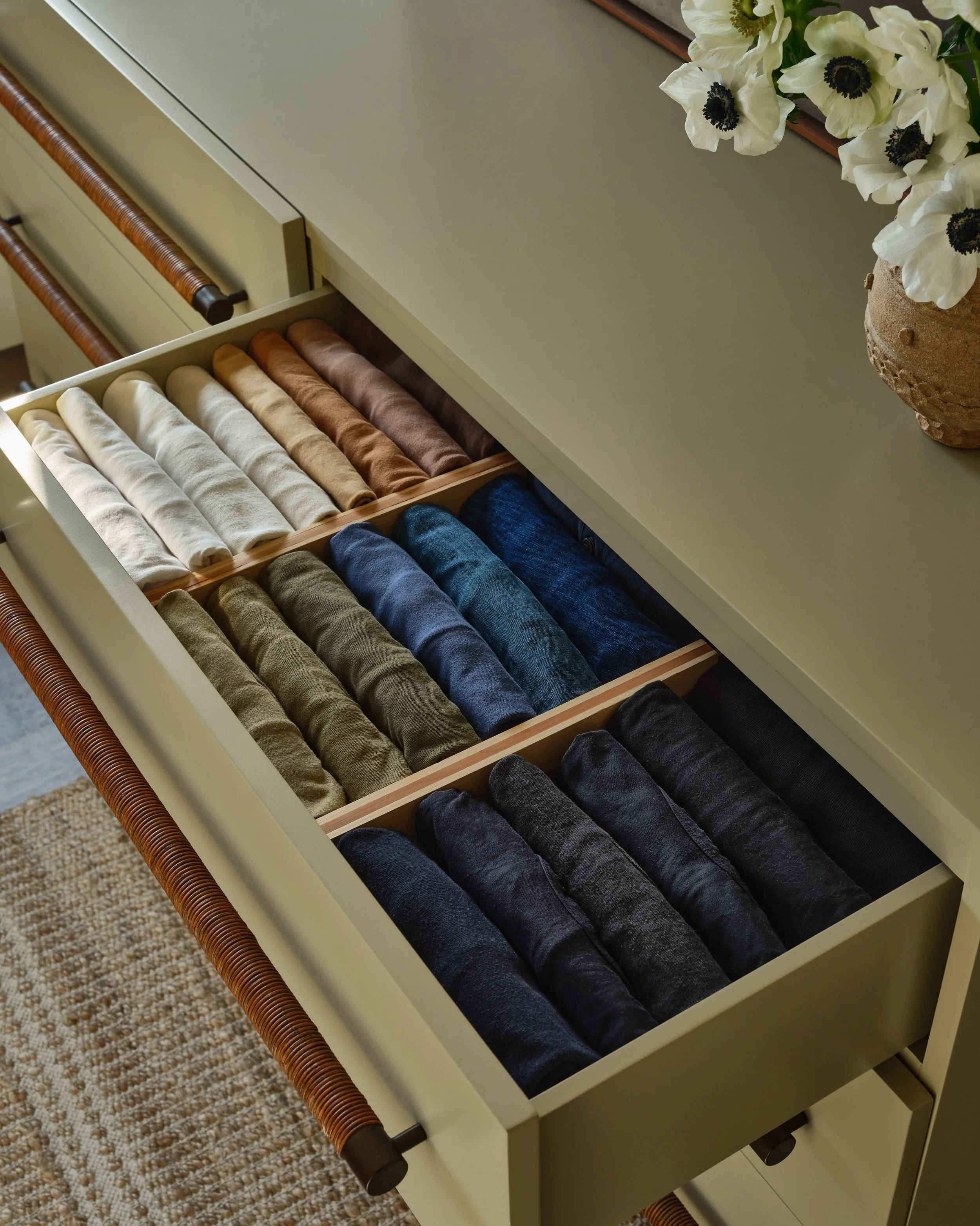 Open drawer with neatly folded socks arranged by color, next to a round vase with white flowers on a beige surface.