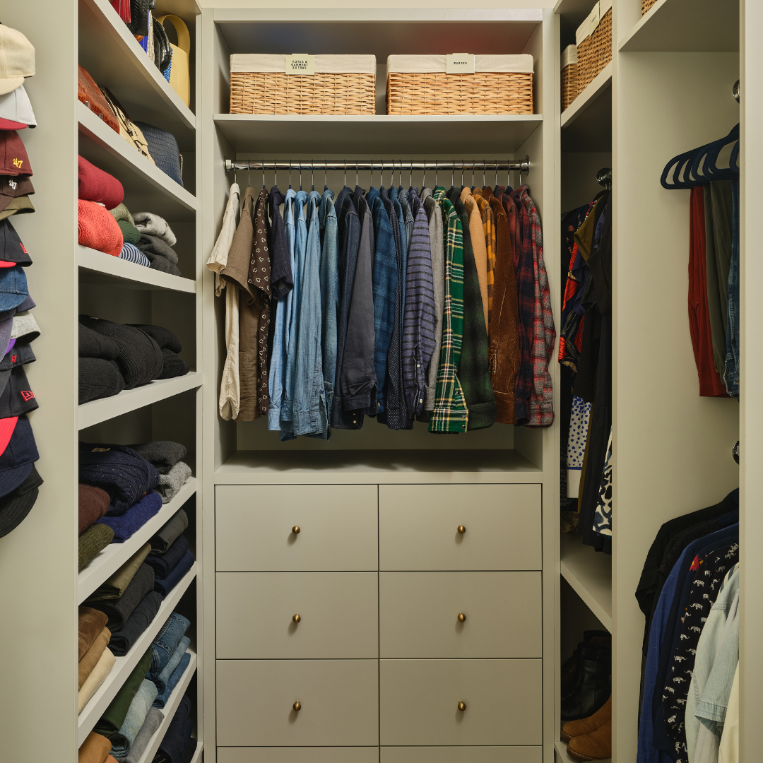 A closet with neatly folded clothes on the left shelves, hanging shirts and jackets in the center, and folded pants on the right, with woven baskets on the top shelves.