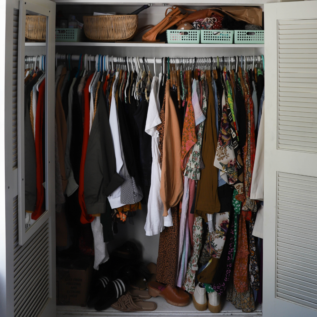 Open closet with hanging clothes of various styles and colors, storage baskets on top shelf, shoes on the bottom, and a partially open louvered door.