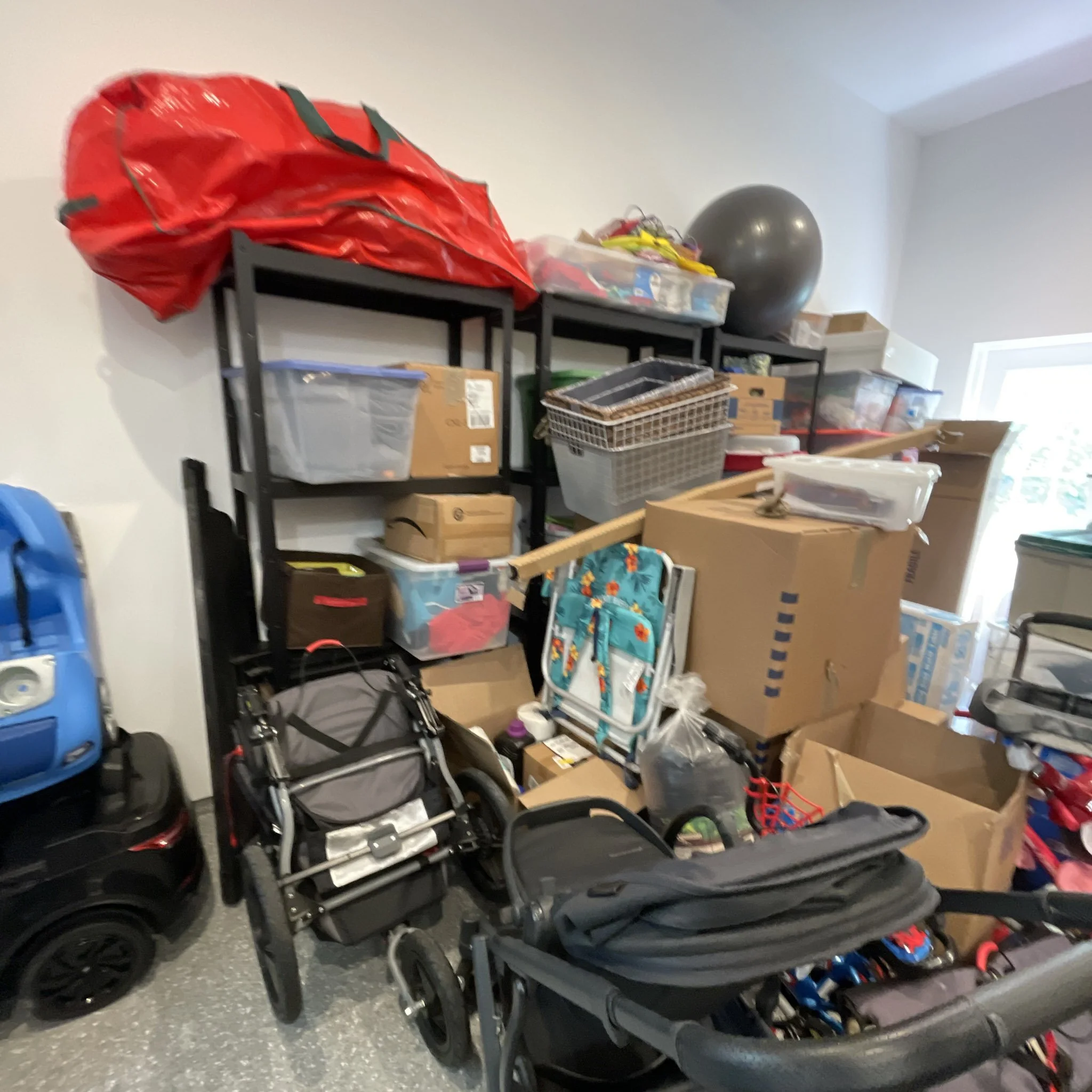 Multiple strollers and boxes stacked on black shelving in a room with white walls and a window.