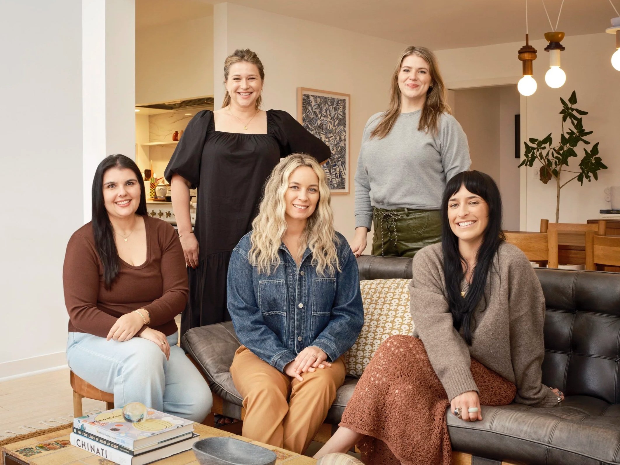 A group of six women posed together in a cozy, modern living room, smiling at the camera. They are sitting and standing around a black leather sofa, with a wooden coffee table in front of them that holds books, a small decorative object, and a sphere. The room has warm lighting, artwork on the walls, and a large potted plant in the background.