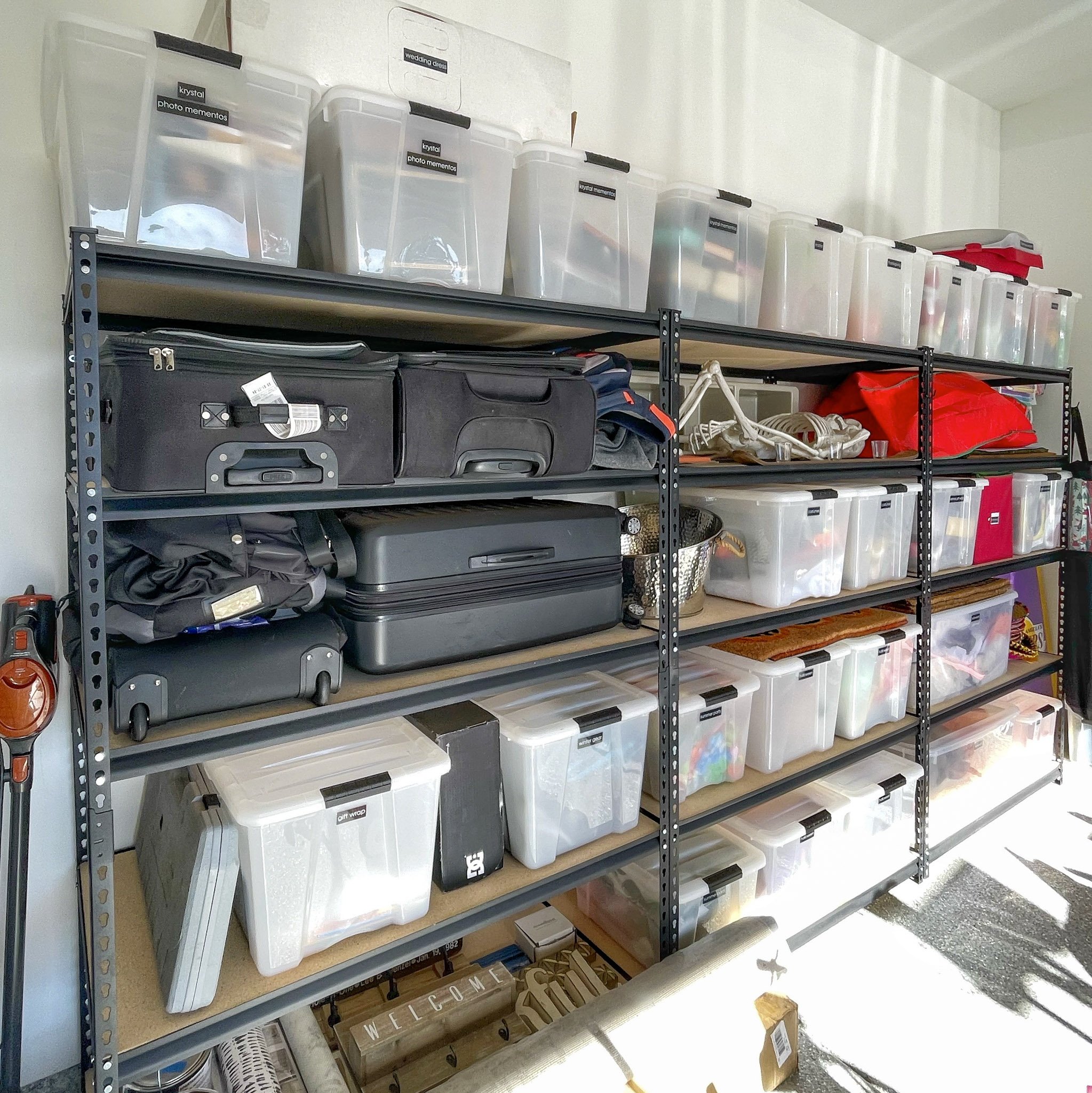 A metal shelving unit holds clear plastic storage bins labeled 'photo mementos', 'wedding dress', and other categories, with various items inside. The bottom shelf contains a black suitcase, a brown wooden sign that says 'WELCOME', and other items, w