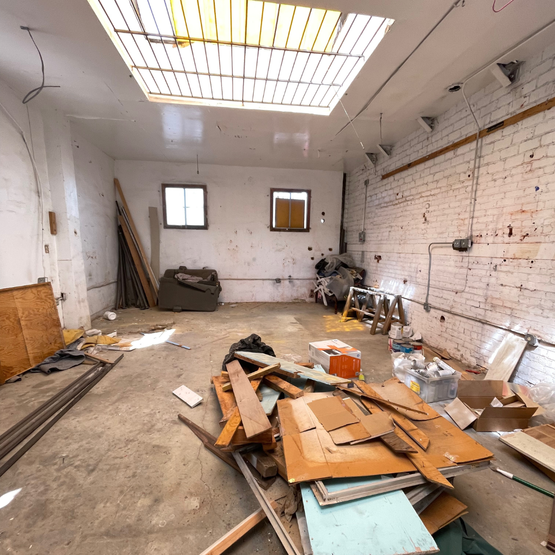 Interior of a cluttered and unfinished room with construction debris, wooden planks, boxes, and tools scattered across the floor. The room has white brick and painted walls, two small windows, and a yellow and white ceiling light panel.