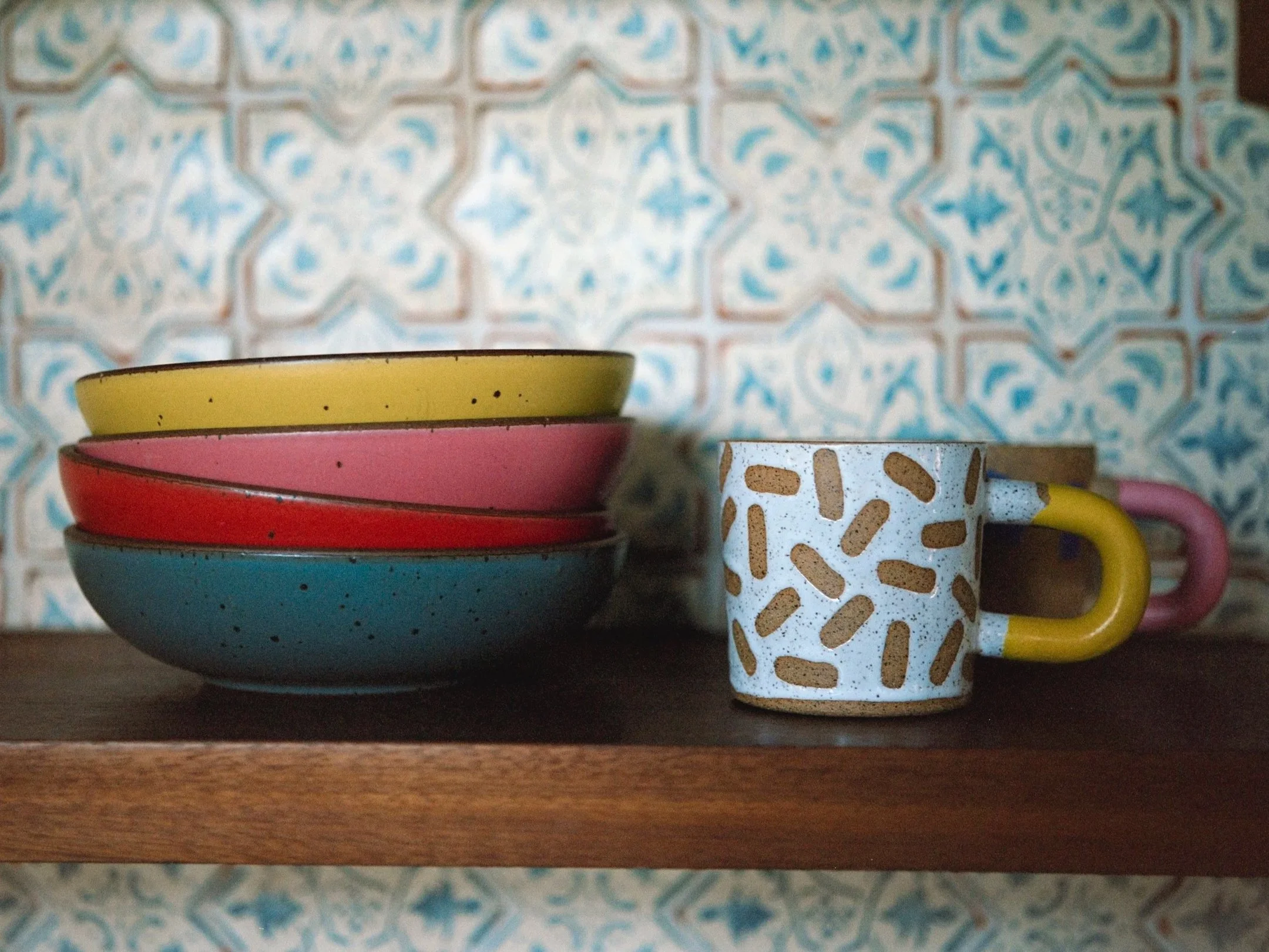 Stacked colorful ceramic bowls and mugs on a wooden shelf against a patterned blue and white tile wall.