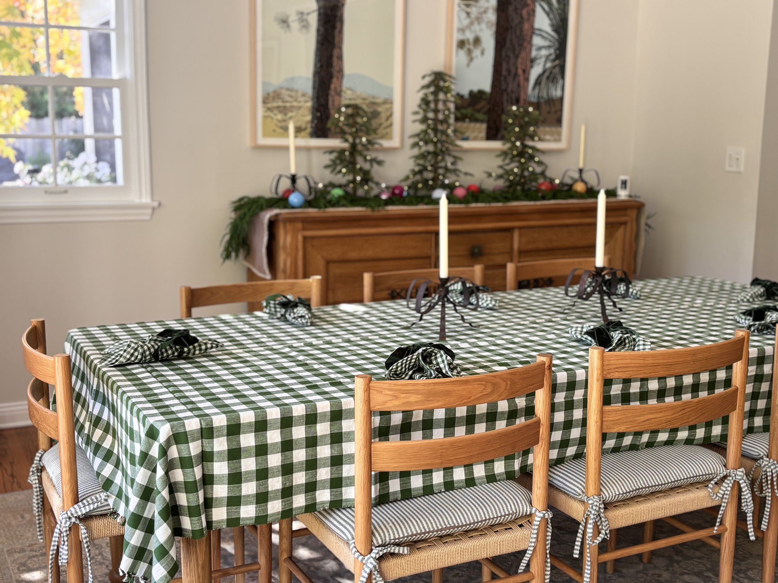 A dining room with a table covered by a green and white checkered tablecloth, surrounded by wooden chairs with striped cushions, decorated for Christmas with napping green garland, colorful ornaments, and candles on a sideboard, and Christmas trees with lights on the wall.