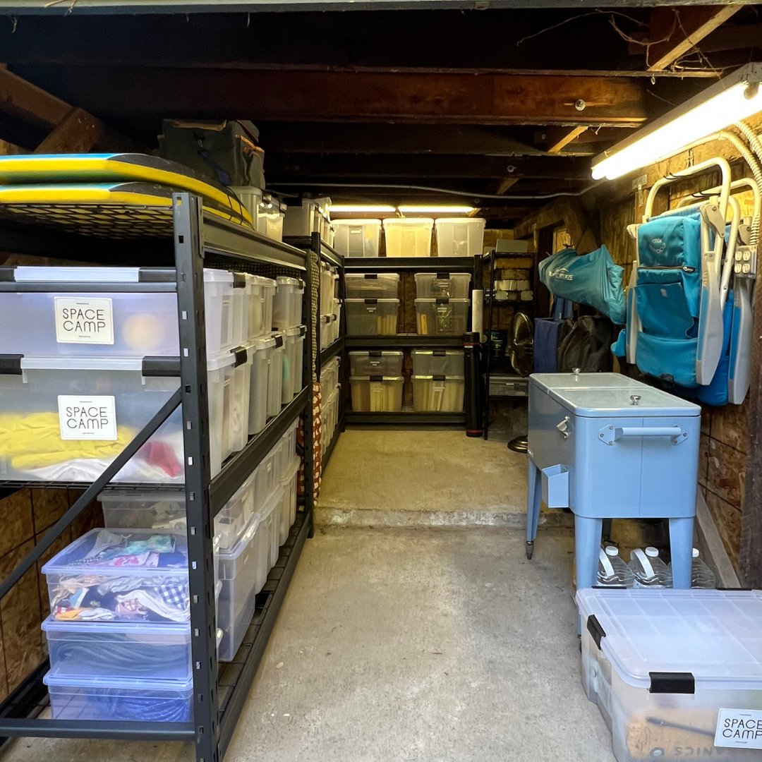 Organized storage shed with shelving units holding plastic containers, backpacks, and storage boxes, a water dispenser, and camping gear.