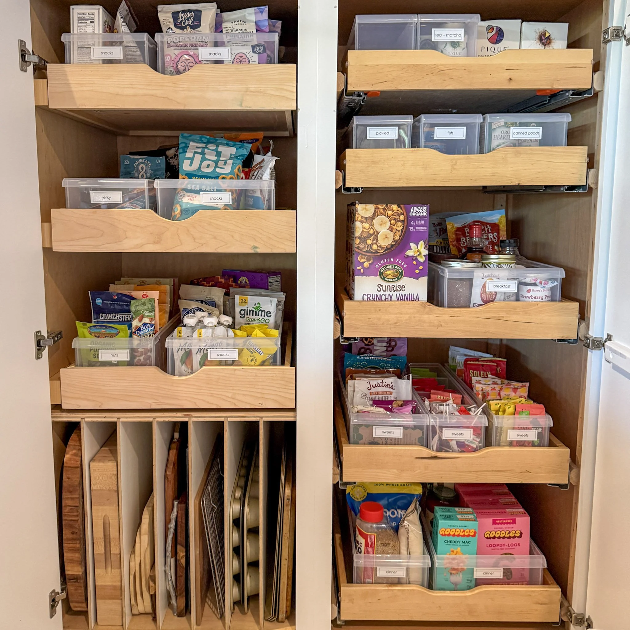 A pantry cabinet with wooden shelves containing organized snacks, grains, and miscellaneous food items in labeled clear plastic bins and containers.