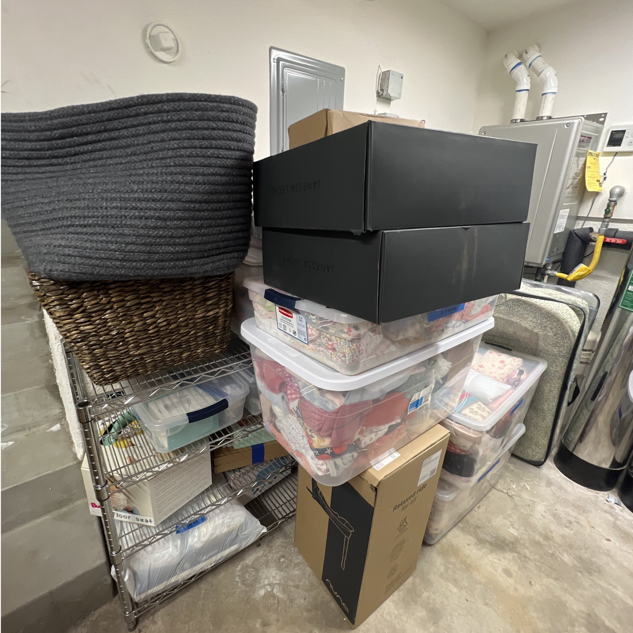 Storage room with a large black basket, metal shelving unit with boxes and containers, and is connected to a utility box and pipes on the wall.