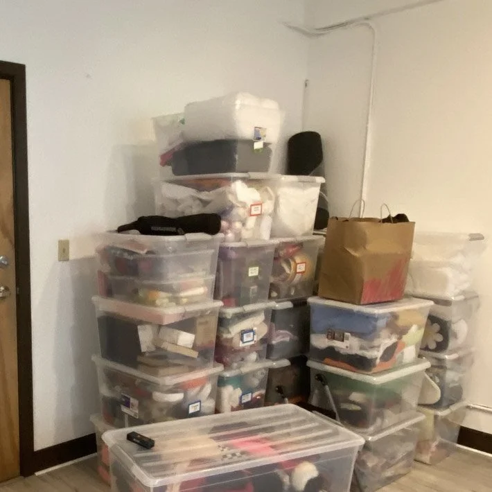 Stack of clear plastic storage bins filled with various craft supplies and materials in a room with a window and door.
