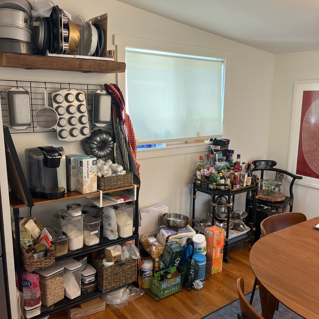 A kitchen corner with shelves filled with baking supplies, a bar cart with liquor bottles, and a dining table with chairs.