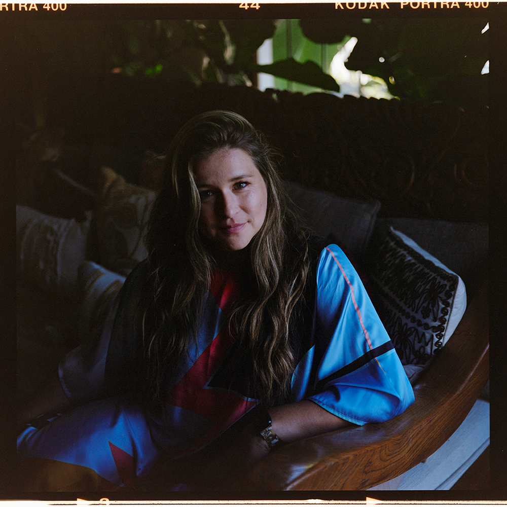 A young woman with long wavy hair, sitting on a dark wood couch, wearing a brightly colored sports jersey. She is indoors with sunlight coming through a window behind her, and decorative pillows are visible behind her.