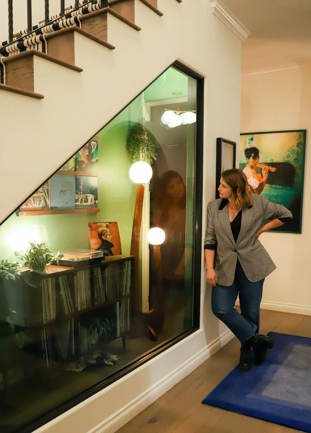 A woman stands next to a large glass window or mirror in a living room, looking at a reflection of herself. There is a painting of a woman playing flute on the wall behind her, and a bookshelf with records and plants below. A large, lit floor lamp with spherical bulbs is also visible.