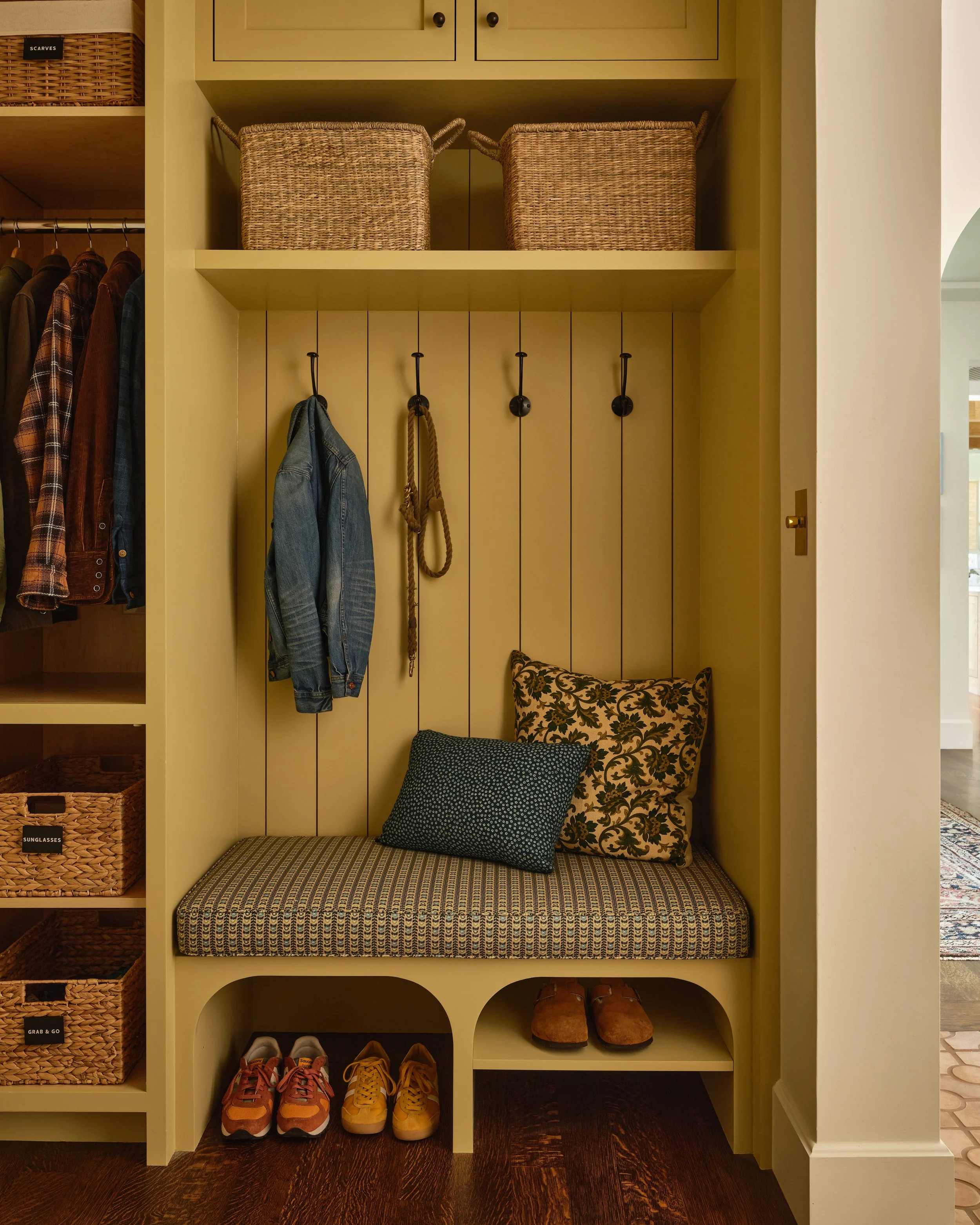 A yellow storage area with hooks holding a denim jacket and a rope, patterned pillows on a bench, shoes on the bottom shelf, and baskets labeled 'Sunglasses' and 'Grab & Go' on the side.